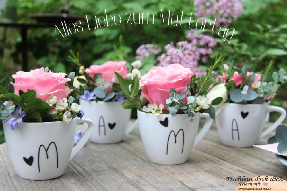 vier Tassen aus weißem Porzellan beschriftet mit den Buchstaben m a m a und kleinen Herzen in schwarz. gefüllt sind die Tassen mit Blumen wie eine große rosafarbene Rose und Eukalyptus