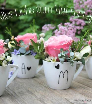 vier Tassen aus weißem Porzellan beschriftet mit den Buchstaben m a m a und kleinen Herzen in schwarz. gefüllt sind die Tassen mit Blumen wie eine große rosafarbene Rose und Eukalyptus