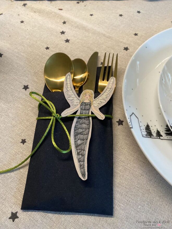 Weihnachtliche Serviettendeko mit goldfarbenem Besteck, schwarzer Serviette und Engel-Anhänger als Tischdeko Idee